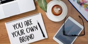 a laptop, plant, coffee and notepad on a desk. The notepad reads "You are your own brand"